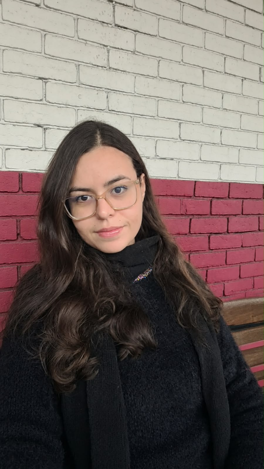 Amanda Queiroz, de óculos e suéter preto, diante de uma parede de tijolos branca e bordô